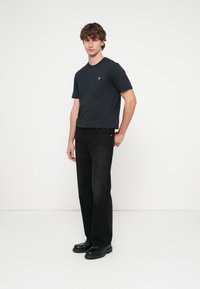 Black cotton t-shirt with a crew neckline and a small yellow logo, paired with black denim jeans and black shoes. Model standing against a plain background.