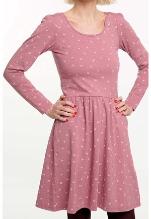 Kobieta w różowej sukience z długim rękawem i małymi białymi kwiatowymi wzorami, z rękami na biodrach i czerwonym lakierem na paznokciach.