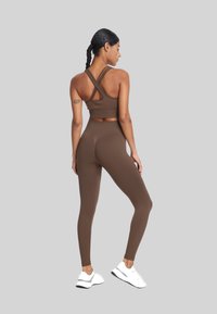 Brązowy stanik sportowy z krzyżującym się tyłem, w zestawieniu z brązowymi legginsami o wysokim stanie. Model nosi białe sneakersy. Gładki, elastyczny materiał.