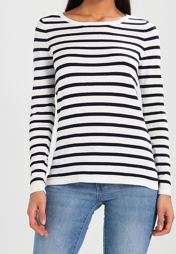 Femme portant une chemise blanche à manches longues avec des rayures horizontales noires et un jean bleu clair, debout les bras détendus le long du corps.