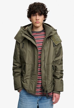 Joven con cabello rizado que lleva una chaqueta con capucha verde oliva sobre una camiseta de rayas rojas y grises y vaqueros azules, con las manos en los bolsillos.
