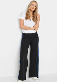 Zwarte rechte broek met blauwe zijstrepen, gecombineerd met een witte T-shirt met korte mouwen. Vloeiende stof en ontspannen pasvorm. Casual schoenen inbegrepen.