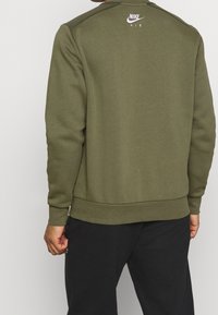 Olivově zelená mikina se širokým výstřihem, žebrovanými manžetami a lemem. Na horní části zad se nachází bílé logo Nike Air. Hladká textura látky.
