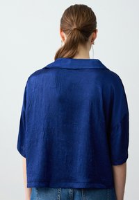Kurze Ärmel, tiefblaue Bluse aus strukturiertem Stoff mit einem klassischen Kragen und einer lockeren Passform. Die Rückansicht zeigt glatte Nähte und eine verkürzte Länge.