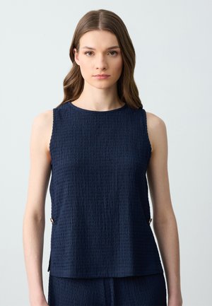 Haut sans manches bleu marine avec un tissu texturé et froissé. Il présente un col rond et des subtils accents latéraux avec des détails en métal.