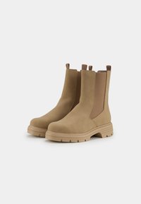 Bottes chelsea en suède beige avec des panneaux latéraux élastiques, des tirants et des semelles en caoutchouc épaisses. Surface texturée avec un design minimaliste.