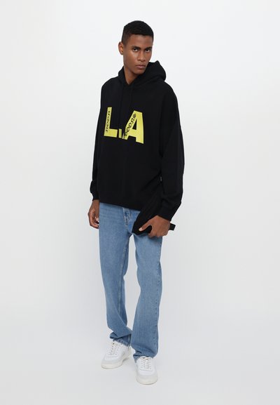 Zwarte hoodie met gele tekst "LA" en "neon" in vet; casual pasvorm. Gepaard met lichtblauwe jeans en witte sneakers. Katoenen materiaal.