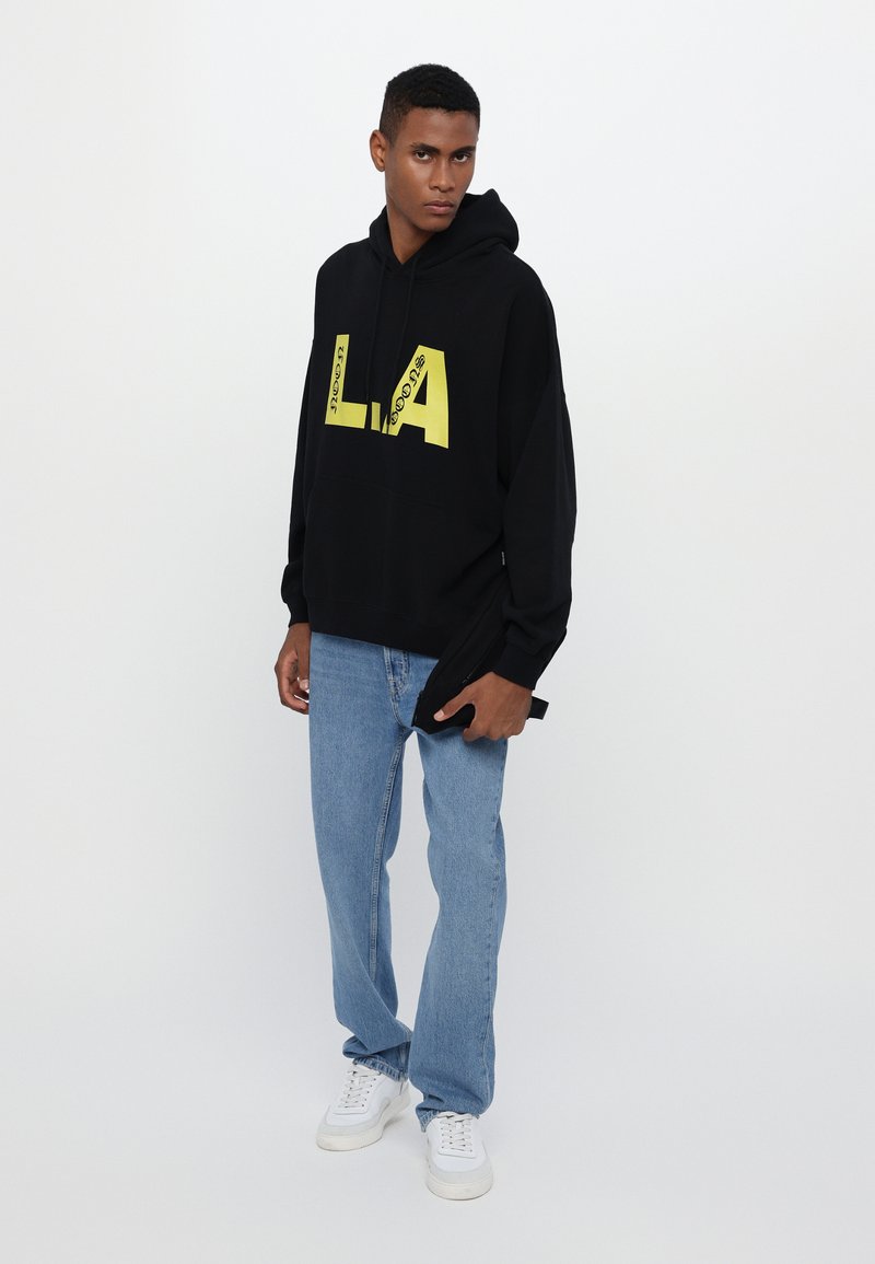 Sweatshirt noir avec le texte jaune "LA" et "neon" en gras ; coupe décontractée. Associé à un jean bleu clair et des baskets blanches. Matière en coton.