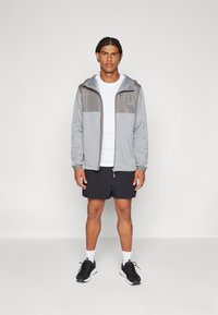 Siva jakna na zatvarač s mješavinom svijetlosive i tamnosive boje, uparena s bijelom majicom i crnim kratkim hlačama. Crne sportske tenisice upotpunjuju izgled.