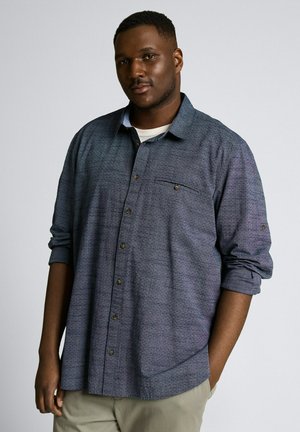 Erwachsener schwarzer Mann mit kurzem Haar, der ein blaues Hemd mit Knöpfen und hochgekrempelten Ärmeln sowie beige Hosen trägt und vor einem schlichten Hintergrund steht.