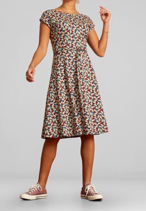 Mujer con un vestido floral de manga corta hasta la rodilla, con cinturón en la cintura y zapatillas de lona rosas, posando frente a un fondo liso.