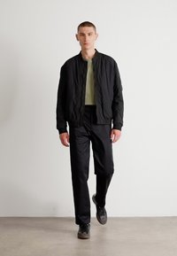Veste bomber noire avec une finition texturée, portée par-dessus une chemise verte, associée à un pantalon noir et des baskets grises, posée devant un fond clair.