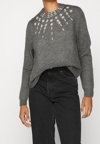 Maglione grigio a maglia con colletto arrotondato, impreziosito da dettagli in perle bianche con un motivo floreale sul petto. Abbinato a jeans neri.