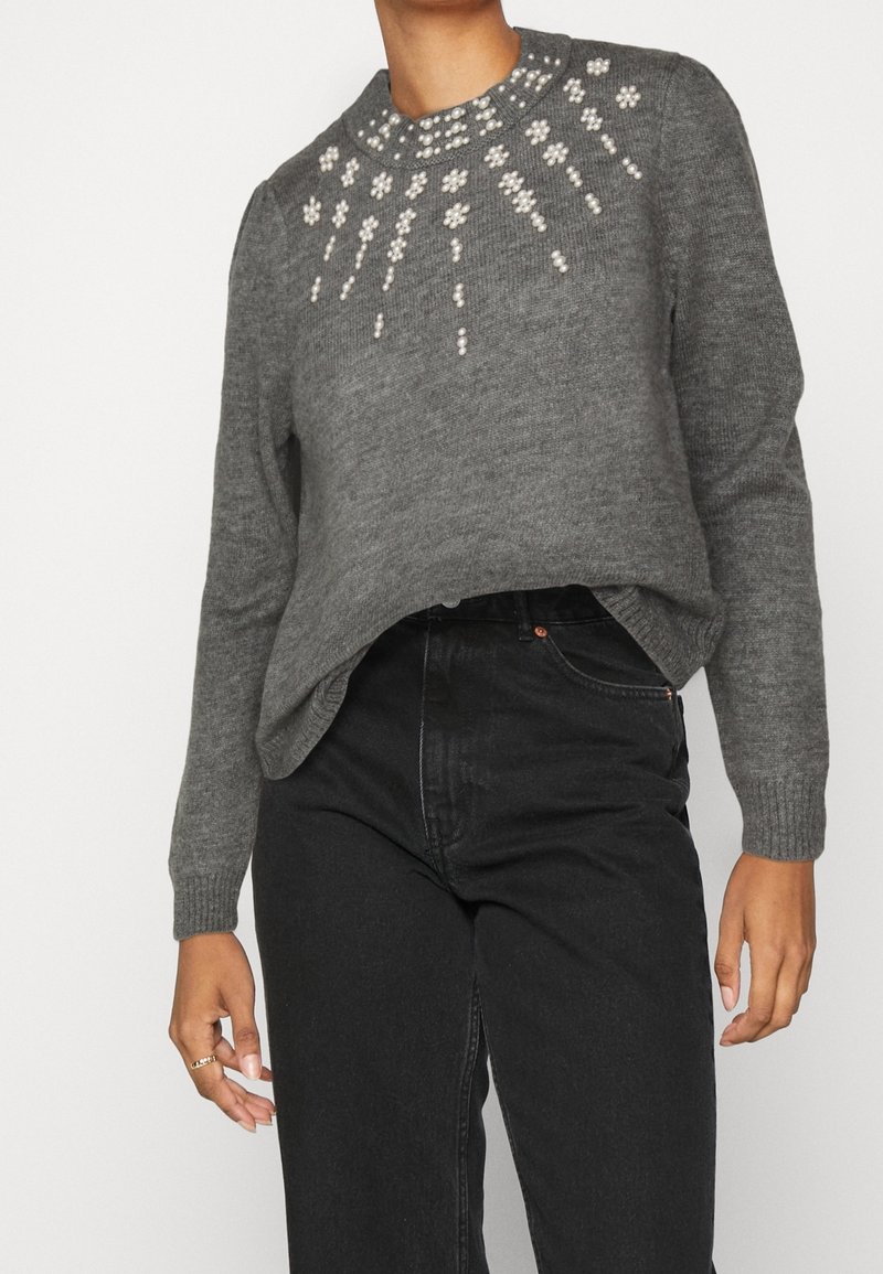 Maglione grigio a maglia con colletto arrotondato, impreziosito da dettagli in perle bianche con un motivo floreale sul petto. Abbinato a jeans neri.