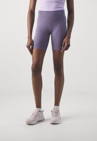 Pantaloni ciclistici viola a vita alta realizzati in materiale elasticizzato, con una texture liscia e un design aderente, abbinati a sneakers sportive rosa chiaro.