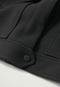 Gros plan d'un tissu noir texturé avec des coutures cousues et un petit bouton rond noir dans le coin.