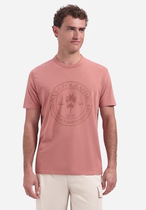 Man draagt een rozenkleurig grafisch T-shirt met palmboom en tekst, gecombineerd met lichtbeige cargoshorts, staand tegen een witte achtergrond.