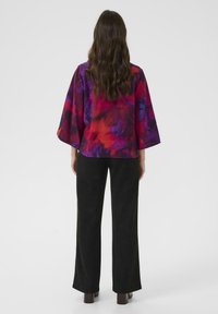 Bluză colorată, cu croială lejeră, prezentând un model abstract vibrant în nuanțe de roșu, violet și albastru, asortată cu pantaloni largi negri și pantofi cu toc bloc maro.