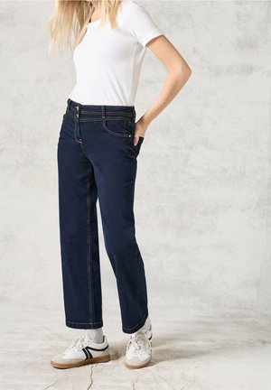 Personne portant un t-shirt blanc, un jean droit bleu foncé, des chaussettes grises et des baskets blanches, debout devant un fond texturé clair.