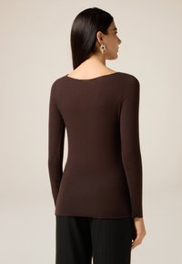 Brown long-sleeve top with a rounded neckline, featuring a smooth texture and fitted design, paired with black striped trousers.
