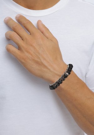 Mandlig hånd iført to sorte perle- og snoede sølvarmbånd, hvilende på brystet over en hvid skjorte.