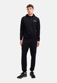 Black hoodie and sweatpants set, cotton material, drawstring hood, ribbed cuffs, and pants. Logo detail on the chest and side of pants. Black sneakers.