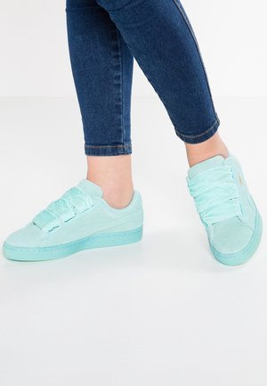 Person iført lyseblå sneakers med tre brede satinremme kombineret med mørkeblå skinny jeans på hvidt gulv.