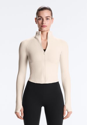 Femme portant une veste beige ajustée avec fermeture éclair, trous pour les pouces et col montant, associée à un pantalon noir taille haute, vue de face.