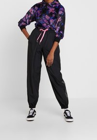 Person klädd i svarta trackpants med rosa snöre, lila och svarts blommig hoodie, och svart-vita sneakers som står framför en vit bakgrund.