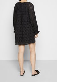 Donna in piedi con le spalle rivolte, indossa un mini vestito lungo nero a fantasia e sandali neri scivolati su un pavimento grigio.