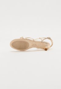 Sandale dorée à fines lanières avec un talon étroit, semelle texturée et petit talon arrière. Caracteristiques : lanières enroulées et finition lisse, design minimaliste.
