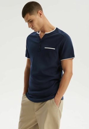 Mężczyzna w granatowej koszulce henley z krótkim rękawem z białą lamówką i beżowych spodniach, ręce w kieszeniach, patrzący w dół na jednolitym tle.