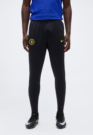 Hombre con pantalones de chándal negros del club de fútbol Chelsea con logotipos amarillos y botas negras sobre un fondo claro.