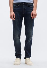 Σ�κούρο μπλε τζιν με εφαρμοστή γραμμή, πέντε τσέπες, διακριτική ξεθώριαση και ραφές. Συνδυασμένο με κρεμ χρώματος αθλητικά παπούτσια.