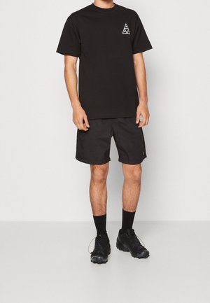 Sort bomulds t-shirt med en trekantet logo på brystet, parret med sorte shorts og sorte sportsko med strukturerede gummisåler.