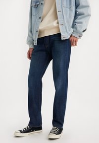 Jeans azul oscuro con corte recto, hechos de tela de mezclilla. Combinados con una chaqueta de mezclilla clara y zapatillas de lona negras. Con costuras de contraste etiquetadas.