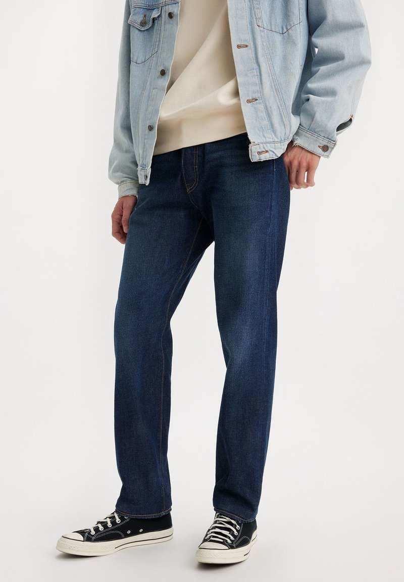 Jeans azul oscuro con corte recto, hechos de tela de mezclilla. Combinados con una chaqueta de mezclilla clara y zapatillas de lona negras. Con costuras de contraste etiquetadas.