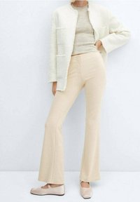 Veste blanche duveteuse avec deux poches, pull beige et pantalon beige évasé. Texture douce et couleurs claires. Chaussures mates de couleur claire.