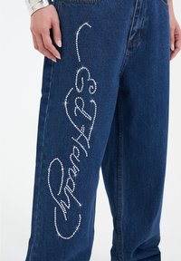 Jeans de mezclilla con corte recto, adornados con un diseño de pedrería a lo largo de la pierna izquierda. La tela es de color azul oscuro con textura y costuras visibles.