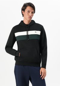Mann med svart hettegenser med hvite og mørkegrønne horisontale striper, og marineblå bukser, står med en hånd i lommen.