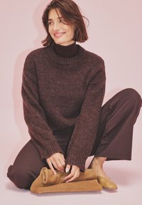 Temno rjav oversized pulover z rebrastim ovratnikom, nošen čez prilegajoč črn uhanski pulover. Model sedi na rjavih semišnih balerinkah, ob njem je zloženo semišno blago.