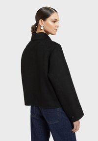 Čierny vlnený kabát so štruktúrovanými ramenami, širokými rukávmi a rovnými líniami. Má klasický golier a gombíkové manžety.