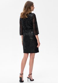 Kobieta w czarnej sukience do kolan z cekinami i przeźroczystymi rękawami, stojąca na białym tle w czarnych sandałach na wysokim obcasie.
