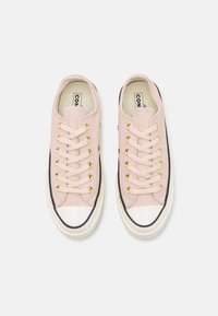 Light pink canvas sneakers with beige laces and gold eyelets. Rubber toe cap features a black trim. Textured surface with minimal detailing.