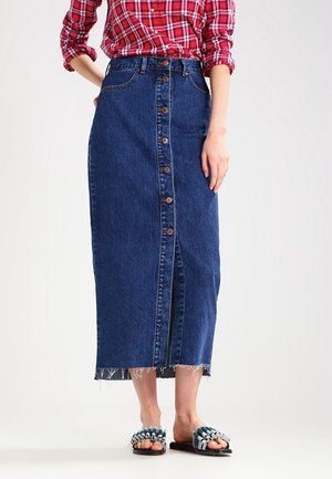 Woman wearing a button-front blue denim midi skirt with frayed hem, red plaid shirt with rolled sleeves, and ruffled slide sandals.
