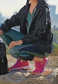 Woman in athleticwear with braided hair crouches on rock outdoors, wearing black jacket, green patterned leggings, and bright pink running shoes.