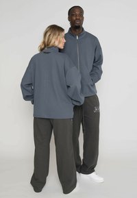 Unisex šedá zipsová bunda s vysokým golierom a štruktúrovanou tkaninou, spárovaná so šedými nohavicami na šnúrku s bočnými zipsami a logovými akcentmi.