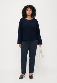 Navy ribbed knit sweater with loose fit and wide sleeves, paired with dark green tapered trousers and brown pointed loafers. Holding a small white bag.