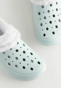 Helles blaues Slipperschuhwerk aus Gummi, mit Belüftungsöffnungen, einem weißen, kuscheligen Innenfutter und einem abgerundeten Zehenbereich.