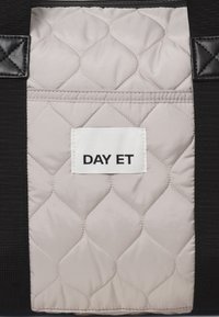 Gepolstertes, weiches beigefarbenes Material mit schwarzen Akzenten; verfügt über ein genähtes weißes Etikett "DAY ET". Auffälliges Diamantmuster und strukturierte Oberfläche.